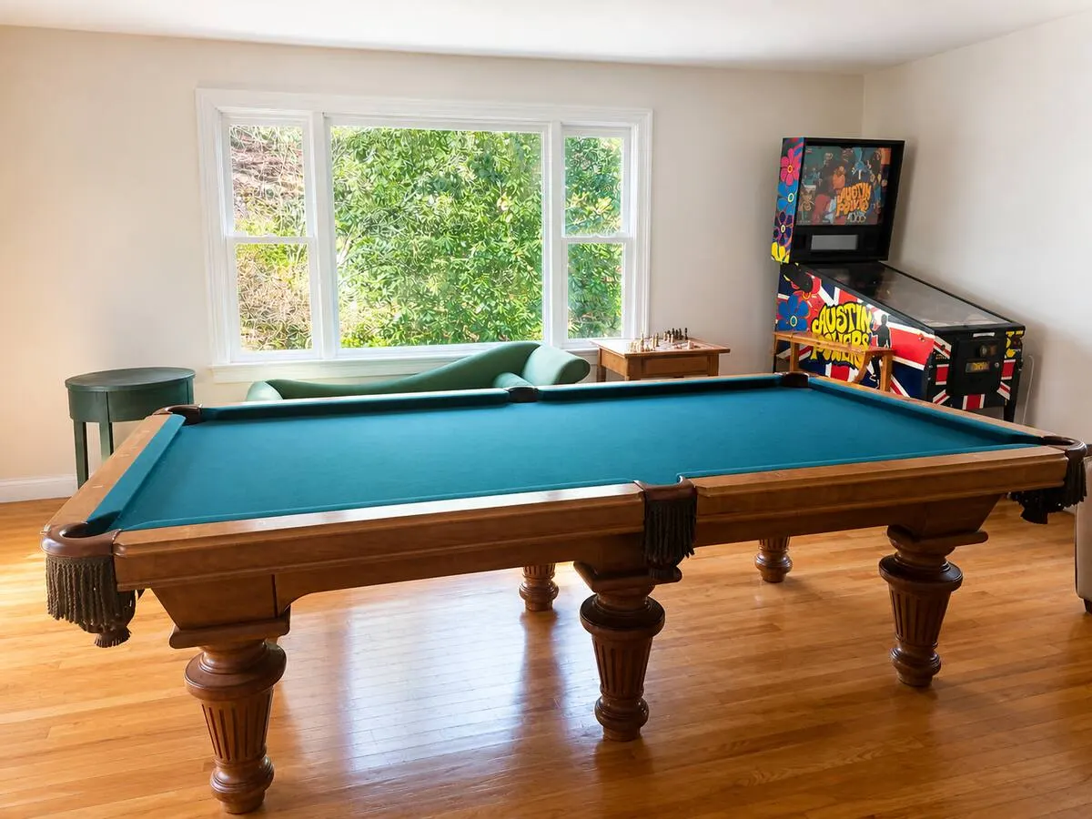 A sunlit room with a pool table and a pinball machine by tall windows